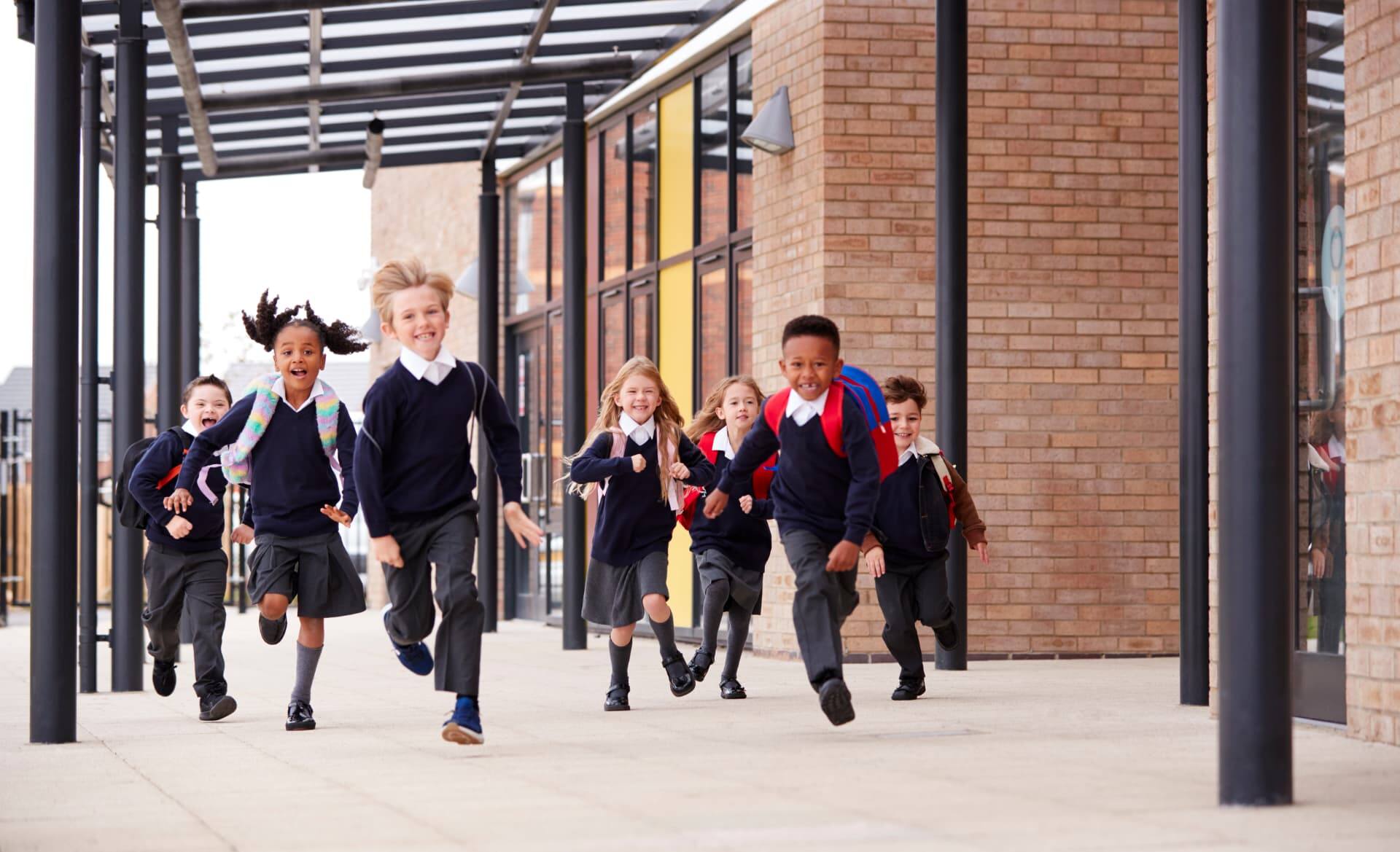 Primary school pupils in uniform running along a covered walkway outside a modern school building
